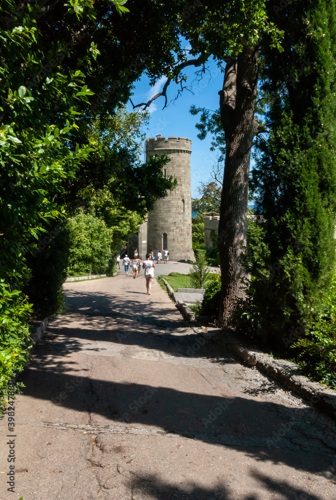 Vorontsov Palace 1848. Tourists walk along a shady, green alley to the watchtowers at the entrance. Alupka, Crimea. Vorontsov Palace 1848. Tourists walk along a shady, green alley to the watchtowers at the entrance. Alupka, Crimea.