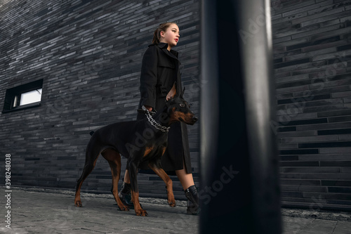 Teen girl walking down the street with her doberman. Great view. Full length of a beautiful young girl in a black cloak. Security and confidence concept