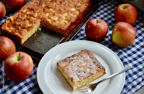One piece of a traditional homemade german apple cake on a white plate with fork, powdered sugar on it, a baking pan behind, all on white and blue squared cotton cloth, apples distributed on it