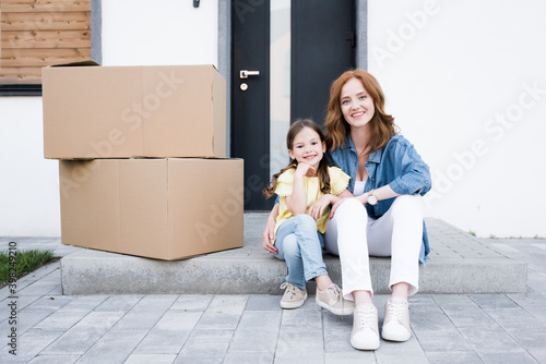 Smiling redhead mother hugging daughter while sitting on doorstep near carton boxes