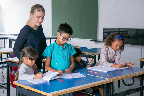 Positive female school teacher watching kids doing their task in class, sitting at desks, drawing and writing in copybooks. Education or back to school concept
