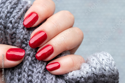 Female hand with gray knitted scarf with beautiful manicure - red glittered nails. Selective focus. Closeup view