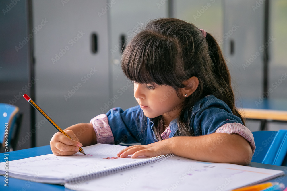 Pensive black haired Latin girl sitting at school desk and drawing in her copybook. Education or back to school concept Pensive black haired Latin girl sitting at school desk and drawing in her copybook. Education or back to school concept