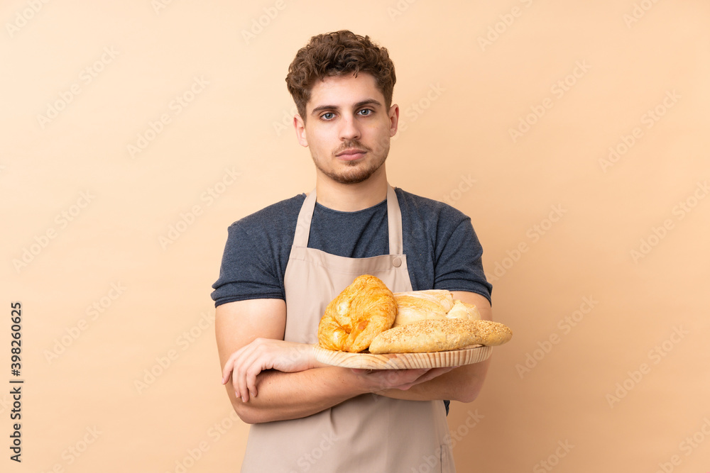 Male baker holding a table with several breads isolated on beige background keeping arms crossed Male baker holding a table with several breads isolated on beige background keeping arms crossed
