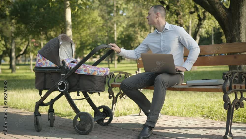 Wide shot of confident busy Caucasian man surfing Internet on laptop and rocking baby carriage sitting on bench in summer park. Portrait of young father combining business and parenthood. Wide shot of confident busy Caucasian man surfing Internet on laptop and rocking baby carriage sitting on bench in summer park. Portrait of young father combining business and parenthood.