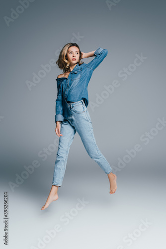 barefoot woman in denim clothes touching hair and looking away while levitating on grey