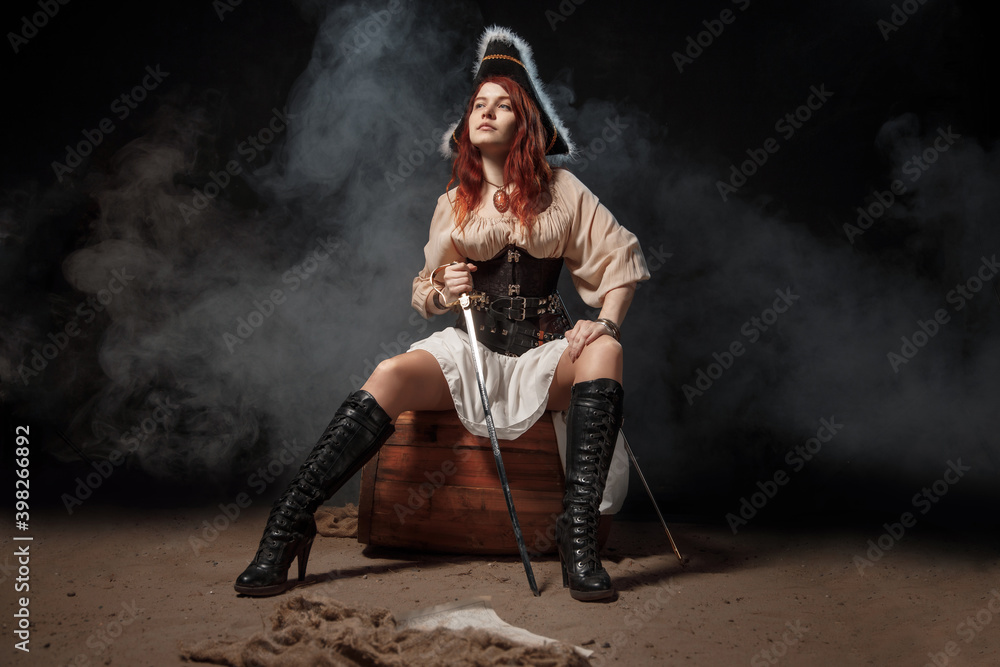 Fototapeta premium Young pirate female with long red hair. Woman is wearing a black corset bustier, tricorn hat , gun belt and armed with a pistol and sword.