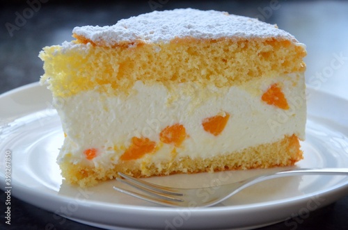 Homemade cream cheese cake with pieces of tangerines in it, powdered sugar on top, sponge cake dough, on a white porcelain plate with fork, blurred background, german and austrian speciality