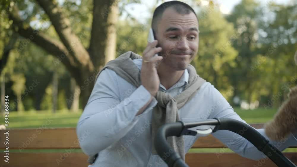 Young desperate father talking on the phone and showing teddy bear to infant in baby stroller. Portrait of tired Caucasian man taking care of baby sitting in summer park. Single parent lifestyle. Young desperate father talking on the phone and showing teddy bear to infant in baby stroller. Portrait of tired Caucasian man taking care of baby sitting in summer park. Single parent lifestyle.