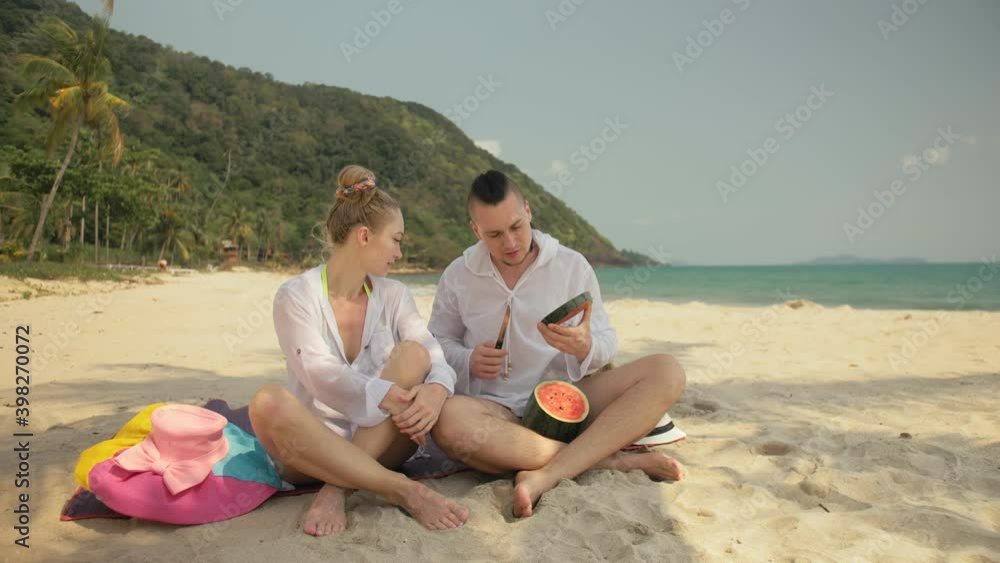 The cheerful love couple holding and eating slices of watermelon on tropical sand beach sea. Romantic lovers two people caucasian spend summer weekend in outdoor. Hat, backpack white shirt beachwear. The cheerful love couple holding and eating slices of watermelon on tropical sand beach sea. Romantic lovers two people caucasian spend summer weekend in outdoor. Hat, backpack white shirt beachwear.
