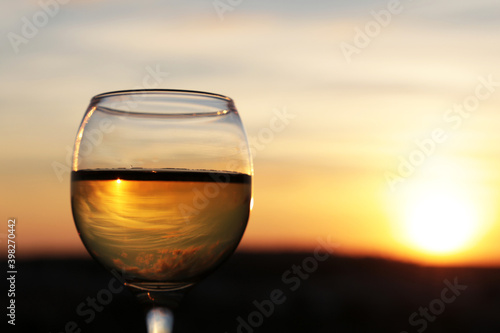 Glass with white wine on beautiful sunset background, sun and sky are reflected in alcohol drink. Concept of celebration, evening party at resort, romantic dinner outdoors