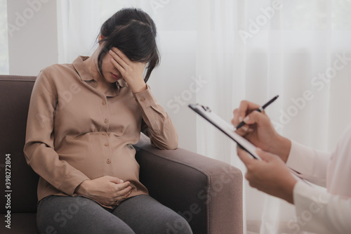 Stressed young asian pregnant woman holding her belly while a psychiatrist notes that the pregnancy is explaining about her unborn child to find out why she worries about motherhood.