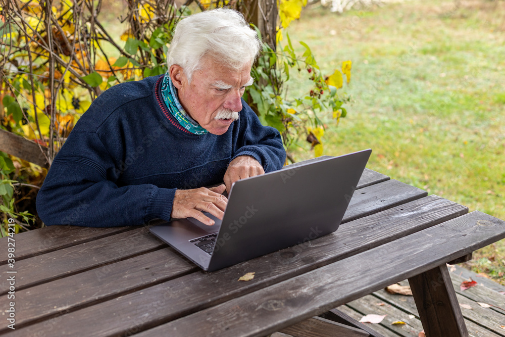 Old man working on laptop in his garden. A senior thoughtful man staring on the screen and making notes.