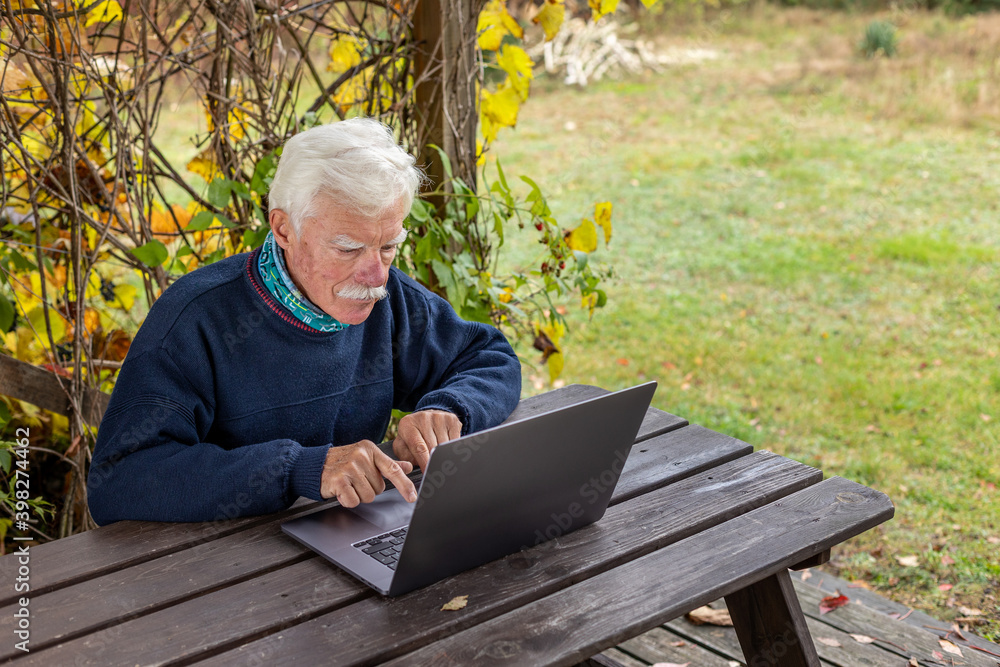 Old man working on laptop in his garden. A senior thoughtful man staring on the screen and making notes.