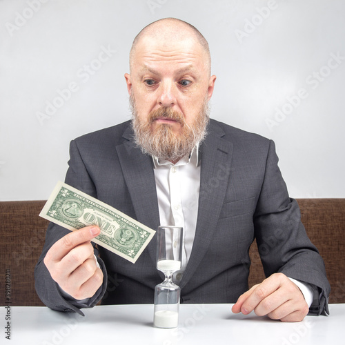 bearded businessman offers payment for work with money against the background of the hourglass. concept of value of time to pay for business.