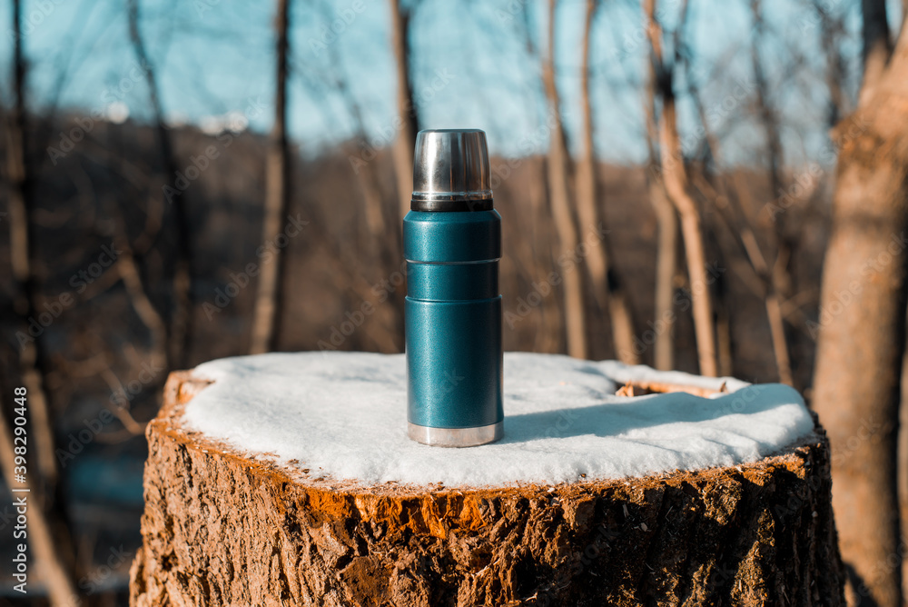 Thermos standing on tree stump covered with snow. Close-up of camping vacuum flask in the winter forest, sunny day. Thermos standing on tree stump covered with snow. Close-up of camping vacuum flask in the winter forest, sunny day.