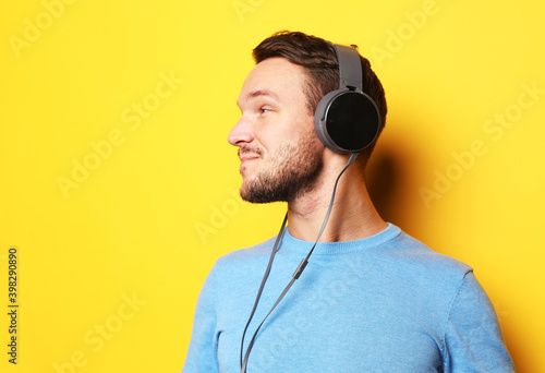 Handsome man wearing blue sweater and headphones and holding mobile phone over yellow background