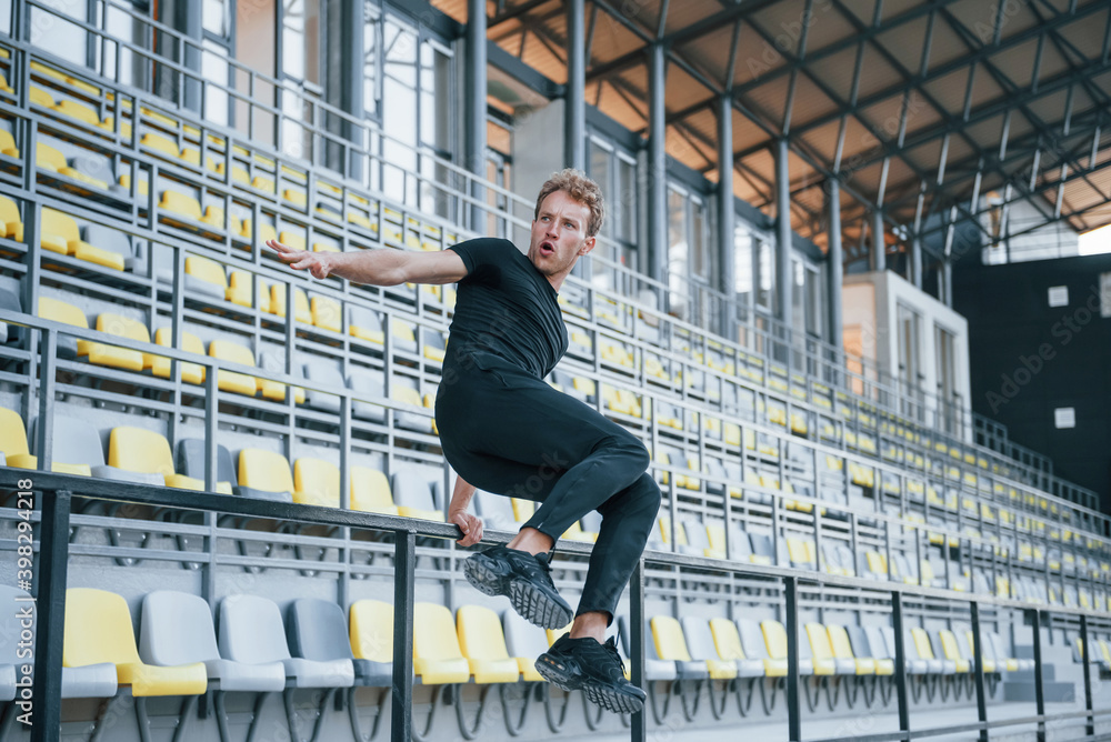 Jumping and doing parkour on the bleachers. Sportive young guy in black shirt and pants outdoors at daytime