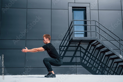 Exercising near building with black wall. Sportive young guy in black shirt and pants outdoors at daytime