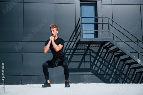 Exercising near building with black wall. Sportive young guy in black shirt and pants outdoors at daytime