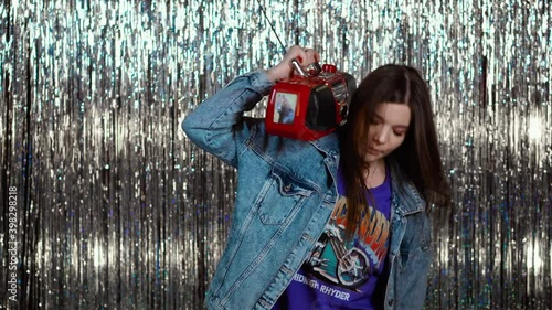 A pretty girl in a denim jacket from the 90s or 80s listens to music and dances. Stylish teenager is holding a retro magnetophone on his shoulder and having fun. photo