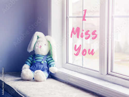 I miss you card with pink letters words text. Cute adorable lonely stuffed toy bunny sitting on a windsill by window. Concept of friends missing each other and loneliness. Children toy left alone. photo