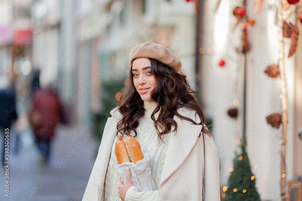 Christmas. Portrait of a young beautiful woman in a beret in a European city. Young woman holds a paper bag with baguettes. 