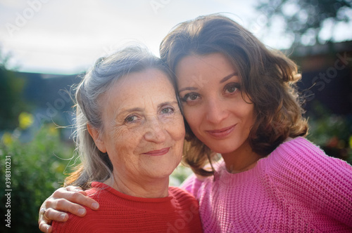 Senior mother with gray hair with her adult daughter looking at the camera in the garden and hugging each other during sunny day outdoors