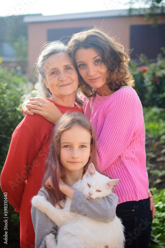 Senior mother with gray hair with her adult daughter and little granddaughter with white cat are looking at the camera in the garden and hugging each other during sunny day outdoors