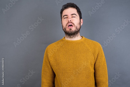 Foto Young handsome Caucasian man wearing yellow sweater against gray wall looking sleepy and tired, exhausted for fatigue and hangover, lazy eyes in the morning