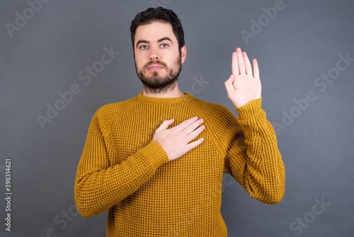 Young handsome Caucasian man wearing yellow sweater against gray wall Swearing with hand on chest and open palm, making a loyalty promise oath photo