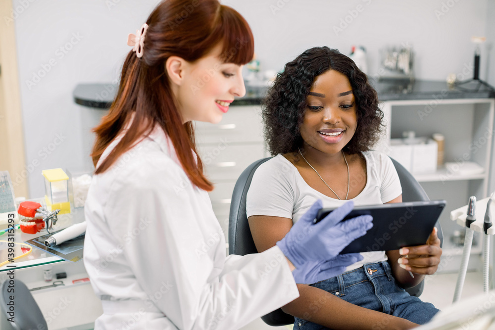 Dentisry concept, healthcare, oral and teeth care. Young beautiful woman dentist discusses the strategy of treatment or shows great results on tablet to young smiling african girl patient Dentisry concept, healthcare, oral and teeth care. Young beautiful woman dentist discusses the strategy of treatment or shows great results on tablet to young smiling african girl patient
