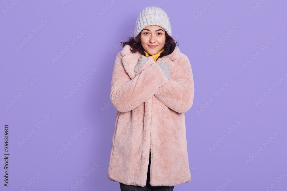 Portrait of young woman in with pale pink faux fur coat and cap, lady keeping hands on her collar and looking directly at camera, needs to warm, standing against lilac wall. Portrait of young woman in with pale pink faux fur coat and cap, lady keeping hands on her collar and looking directly at camera, needs to warm, standing against lilac wall.