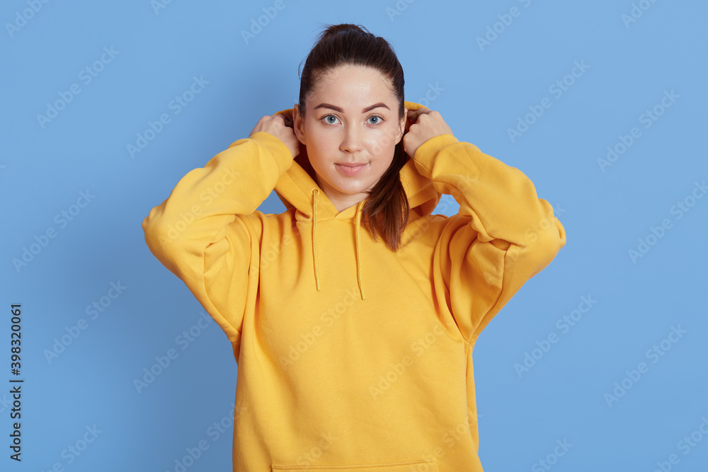 Dark haired woman wearing white hoodie posing at isolated over blue background, looks at camera, has serious facial expression, has pleasant look. keeping hands on hood. Dark haired woman wearing white hoodie posing at isolated over blue background, looks at camera, has serious facial expression, has pleasant look. keeping hands on hood.