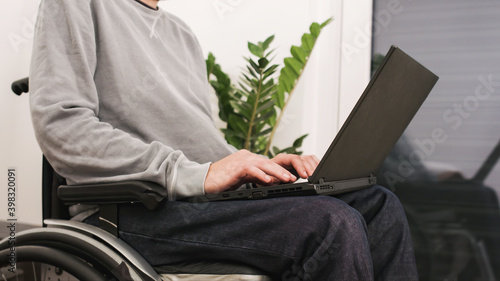 Young man in wheelchair with laptop working remotely from home, using internet and looking at screen.