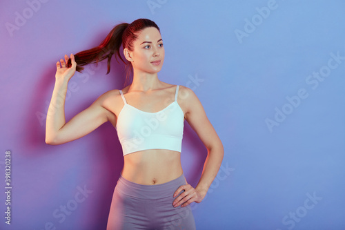 Confident active healthy woman in sports clothing with one hand on hip posing isolated over clor background, tocghing her ponytail and looking away, copy space for advertisement. photo