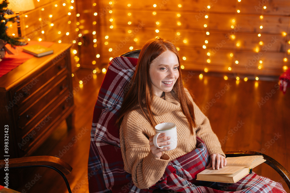 Portrait of relaxed redhead young woman holding cup with hot coffee and reading book while sitting at rocking armchair with knitted plaid, background of wall decorated Christmas sparkling lights.