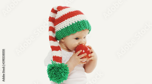 Portrait of baby in knitted gnome winter hat biting red apple over a white background