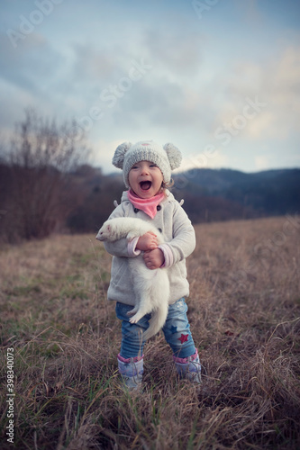 A little girl is outside with her pet. Cute baby girl holding a ferret in a hand. The child and his living furry pet. Autumn in the countryside. Friendship of children and animals. Walking with a