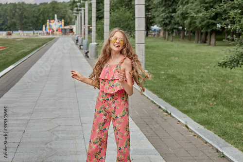 A teenage girl laughs in a pink fashion jumpsuit in a summer Park. Girl in sunglasses. Long blonde wavy hair.