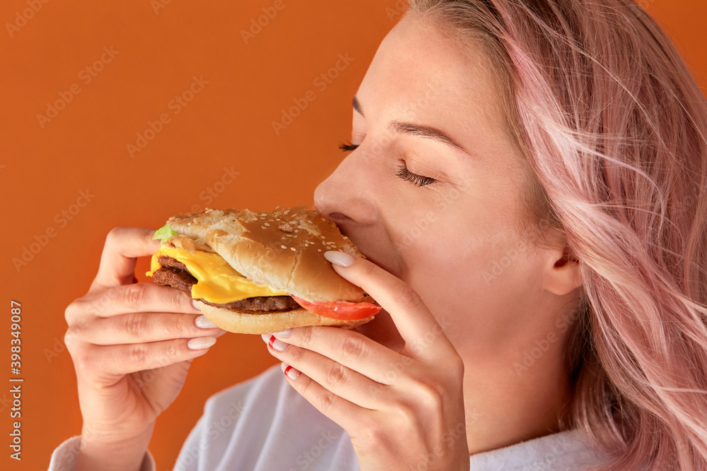 A young woman with pink hair eats fast food with relish. Orange background. The concept of junk food. A young woman with pink hair eats fast food with relish. Orange background. The concept of junk food.