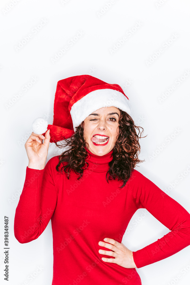 A funny portrait of woman with a Santa hat in Christmas. She is wearing a red sweater and she is looking at the camera with her tongue out. Christmas portrait concept.