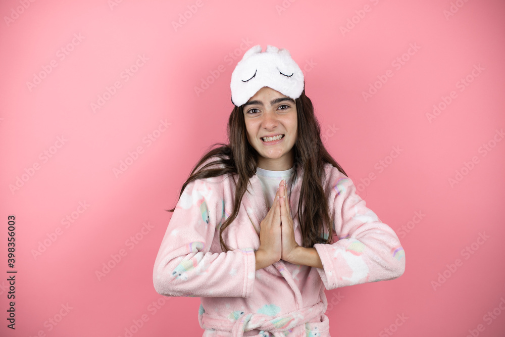 Pretty girl wearing pajamas and sleep mask over pink background begging and praying with hands together with hope expression on face very emotional and worried Pretty girl wearing pajamas and sleep mask over pink background begging and praying with hands together with hope expression on face very emotional and worried