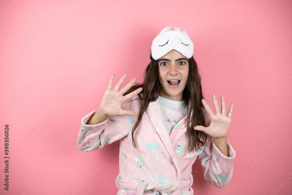 Pretty girl wearing pajamas and sleep mask over pink background afraid and terrified with fear expression stop gesture with hands, shouting in shock. Panic concept. Pretty girl wearing pajamas and sleep mask over pink background afraid and terrified with fear expression stop gesture with hands, shouting in shock. Panic concept.