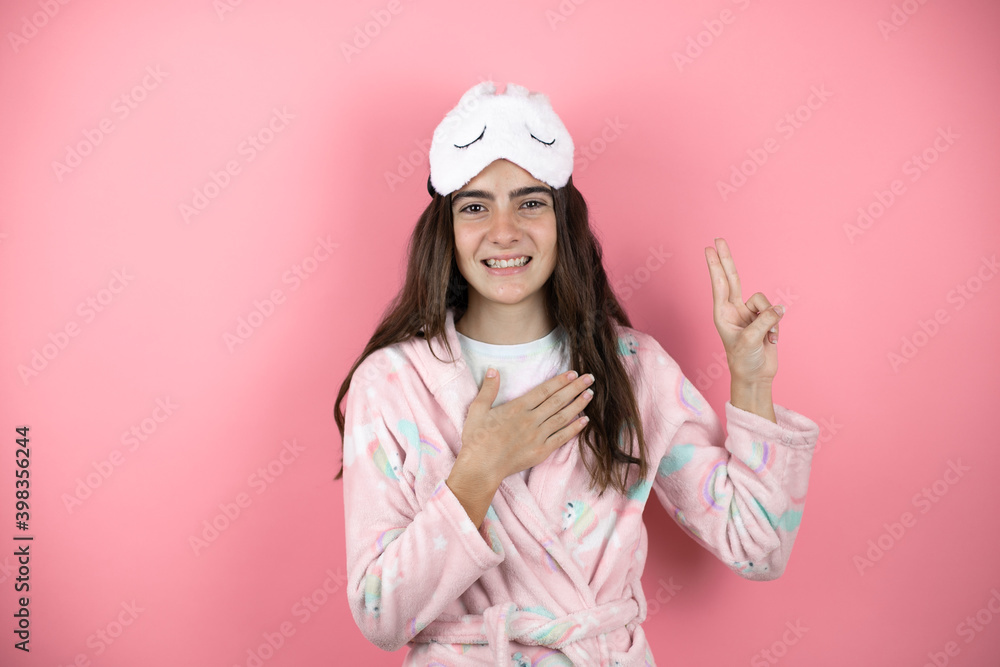 Pretty girl wearing pajamas and sleep mask over pink background smiling swearing with hand on chest and fingers up, making a loyalty promise oath Pretty girl wearing pajamas and sleep mask over pink background smiling swearing with hand on chest and fingers up, making a loyalty promise oath