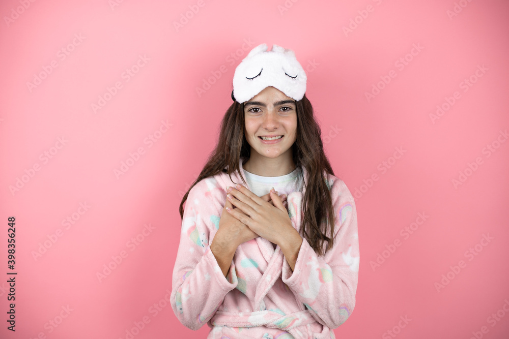 Pretty girl wearing pajamas and sleep mask over pink background smiling with her hands on her chest and grateful gesture on her face. Pretty girl wearing pajamas and sleep mask over pink background smiling with her hands on her chest and grateful gesture on her face.