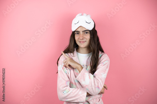 Pretty girl wearing pajamas and sleep mask over pink background smiling happy pointing with hand and finger to the side © Irene