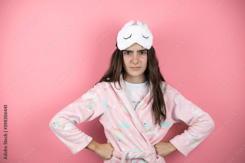 Pretty girl wearing pajamas and sleep mask over pink background skeptic and nervous, disapproving expression on face with arms in waist Pretty girl wearing pajamas and sleep mask over pink background skeptic and nervous, disapproving expression on face with arms in waist