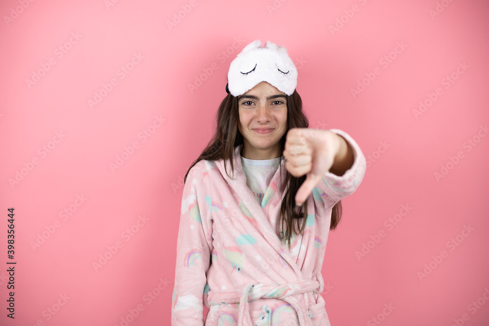 Pretty girl wearing pajamas and sleep mask over pink background with angry face, negative sign showing dislike with thumb down Pretty girl wearing pajamas and sleep mask over pink background with angry face, negative sign showing dislike with thumb down
