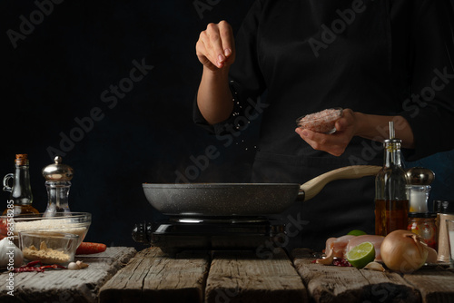 Chef pours sea salt into pan with frying chicken fillet. Backstage of cooking traditional Indian chicken curry on dark blue background. Frozen motion. Concept cooking process. Frozen motion.
